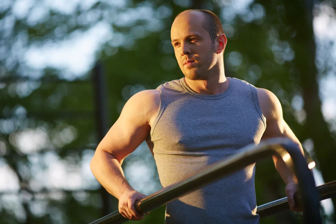 Fit man in a sleeveless shirt using parallel bars outdoors, symbolizing muscle health and physical performance benefits associated with peptide therapy.