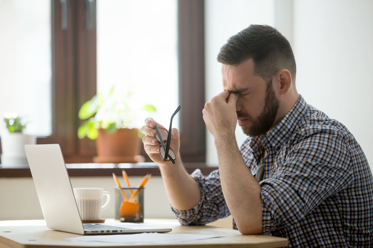 Tired man sitting at a desk with his laptop, holding his glasses and rubbing his eyes in frustration, possibly experiencing symptoms of low testosterone like fatigue or brain fog.